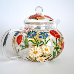 Può includere: Una teiera di vetro trasparente con un motivo floreale dipinto sul lato. I fiori sono rossi, blu, bianchi e verdi. La teiera ha un coperchio e un manico.