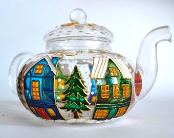 May include: A clear glass teapot featuring a hand-painted winter village scene with a blue house, a green house, and a central evergreen tree. The lid and body are decorated with gold stars and white snow accents, with a curved handle and spout.