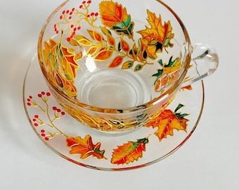 May include: A clear glass teacup and saucer set featuring hand-painted autumn motifs. The design includes vibrant orange, yellow, and green maple and oak leaves, accented with delicate clusters of small red berries outlined in gold paint.