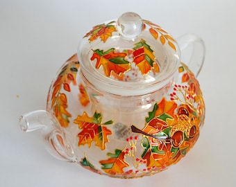May include: A clear glass teapot featuring a hand-painted design of vibrant autumn leaves in shades of orange, red, yellow, and green. The teapot includes a matching glass lid, a curved handle, and a spout, all decorated with painted acorns and berries.