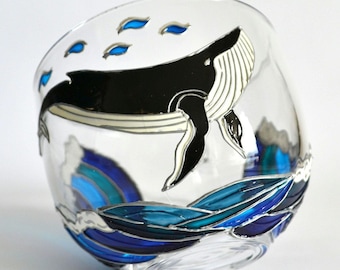 May include: A clear glass bowl featuring a hand-painted black and white whale swimming above stylized blue and dark blue ocean waves. Small blue accents are painted above the whale, creating a marine-themed design on the curved glass surface.