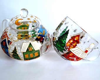 May include: A clear glass teapot and matching teacup featuring hand-painted winter village scenes. The designs include colorful houses with snow-covered roofs, evergreen trees, and a night sky with a golden crescent moon and scattered stars.