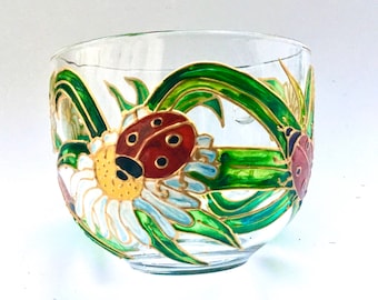 May include: A clear glass cup featuring a hand-painted design of red ladybugs with black spots resting on white daisy flowers with yellow centers, surrounded by vibrant green leaves and stems outlined in gold paint against the transparent glass.
