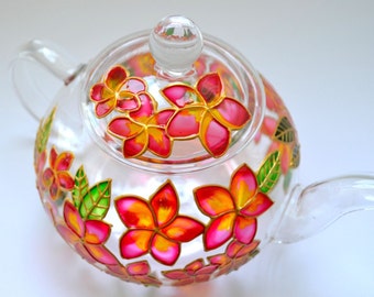 May include: A clear glass teapot featuring a hand-painted floral design with vibrant pink, orange, and yellow petals outlined in gold. The teapot includes a matching glass lid with a knob handle, a curved spout, and a handle, all decorated with green leaves.
