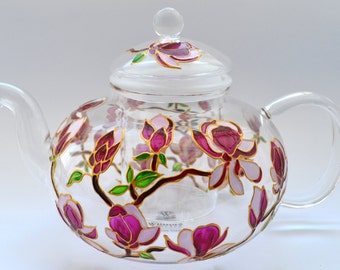 May include: A clear glass teapot featuring a hand-painted floral design with vibrant magenta and light pink magnolia blossoms, green leaves, and brown branches. The teapot includes a matching glass lid with a knob and a clear glass infuser insert.