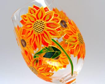 May include: A clear glass wine glass decorated with hand-painted sunflowers featuring vibrant orange and yellow petals, dark brown centers, and green leaves. The floral design wraps around the bowl of the glass with bold outlines.