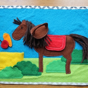 May include: A felt play mat with a brown horse with a red saddle, a yellow bucket, a red apple, and green bushes on a blue background.