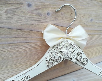 May include: White wooden hanger with a cream satin bow and a pearl embellishment. The hanger is decorated with a seashell, starfish, and intricate designs. The hanger is personalized with the name "Jason" and the date "10.7."