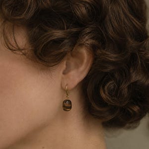 May include: A gold hoop earring featuring a single, smooth, oval-shaped tiger's eye gemstone pendant. The gemstone displays characteristic brown and golden-yellow banded patterns with a polished, reflective surface, suspended from a thin gold wire hoop.