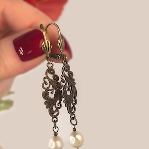 Puede incluir: Pendientes ornamentados de estilo antiguo con acabado bronce. Los pendientes presentan un delicado diseño de filigrana arremolinada y están acentuados con perlas en forma de lágrima. Una mano con esmalte de uñas rojo sostiene los pendientes.