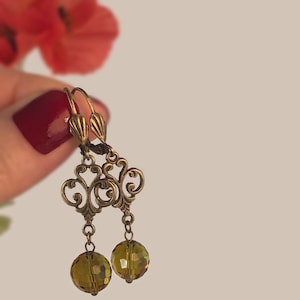 Puede incluir: Un par de pendientes de estilo vintage en tono latón, con intrincados diseños de filigrana. Cada pendiente presenta una cuenta de cristal facetada de color verde oliva. Sobre un fondo neutro, con una flor roja borrosa en la esquina superior izquierda.