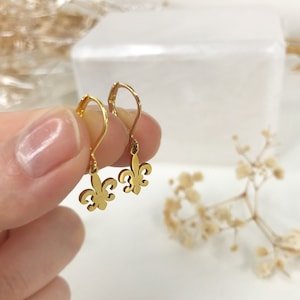 May include: A pair of gold-toned dangle earrings featuring a fleur-de-lis design. The earrings have a simple hook closure.