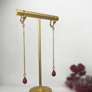 Könnte beinhalten: Ein Paar goldfarbene Ohrhänger mit Kettenmuster. Jeder Ohrring hat eine kleine, tropfenförmige, dunkelrote Perle am Ende der Kette. Die Ohrringe werden auf einem goldfarbenen Ständer präsentiert.