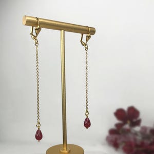 Könnte beinhalten: Goldfarbene Ohrringe mit einer langen Kette und einem tropfenförmigen, dunkelroten Edelstein. Die Ohrringe werden auf einem goldenen Schmuckständer präsentiert. Der Ständer hat einen runden Sockel und eine horizontale Stange.