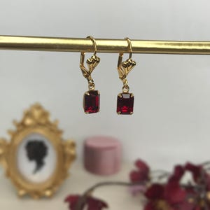 May include: A pair of gold-tone leverback earrings featuring rectangular, deep red faceted stones. The earrings hang from a thin gold-coloured horizontal bar. Each earring has a small, decorative fan-shaped metal detail above the red stone.