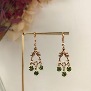 May include: A pair of gold-coloured chandelier earrings with three green gemstone drops. The earrings have an ornate, filigree design with a lever back closure. The earrings are displayed on a gold-coloured stand.