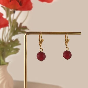 Könnte beinhalten: Ein Paar goldfarbene Klappbügel-Ohrringe mit facettierten, runden, roten Perlen. Die Ohrringe werden auf einem goldfarbenen Ständer präsentiert, mit roten Blumen im Hintergrund.