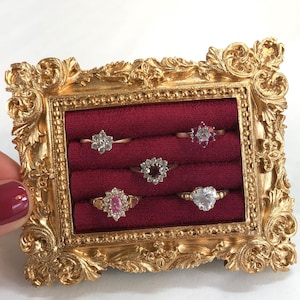 May include: A rectangular gold-toned ornate frame featuring a deep red velvet interior with five slots holding various rings. The rings include a floral-shaped diamond ring, a pink gemstone ring, and a heart-shaped stone ring set in metal bands.