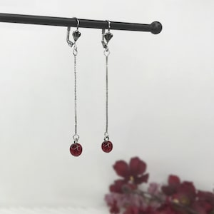 May include: A pair of silver-toned drop earrings featuring a thin box chain suspended from a leverback closure. Each earring terminates in a small, round, translucent red bead that hangs freely at the bottom of the delicate metallic chain structure.