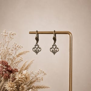 May include: A pair of antique bronze-toned metal earrings featuring a symmetrical, openwork filigree design with curved scrollwork patterns. The earrings are suspended from lever-back closures and hang from a thin, vertical gold-toned metal stand.