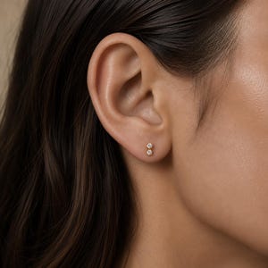 May include: A gold stud earring featuring two small, clear, round-cut stones set vertically in a delicate metal frame. The earring is positioned in the lower earlobe, showcasing a minimalist design with a polished finish and secure backing.