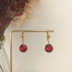 May include: A pair of gold-toned leverback earrings featuring round, faceted red stones. The earrings are suspended from a thin, gold-colored metal display stand. Each earring consists of a circular red stone set in a gold-toned metal bezel.