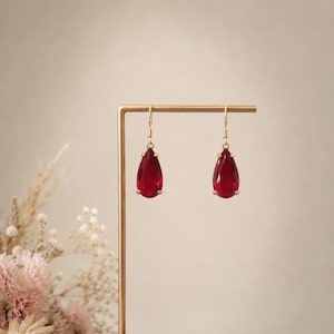 May include: A pair of drop earrings featuring deep red, pear-shaped faceted stones suspended from thin gold-toned metal hooks. Each stone is secured by four small prongs, creating a classic teardrop silhouette that hangs vertically from the metal wire.