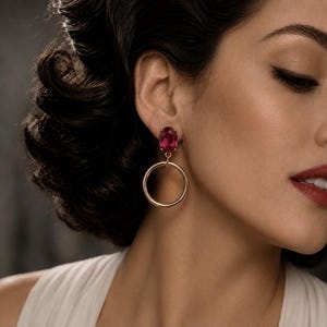 May include: A gold-toned earring featuring a faceted oval-shaped deep pink gemstone stud connected to a smooth, circular gold-toned hoop pendant. The earring is worn on the earlobe, showcasing the contrast between the vibrant pink stone and the metallic gold finish of the circular hoop design.