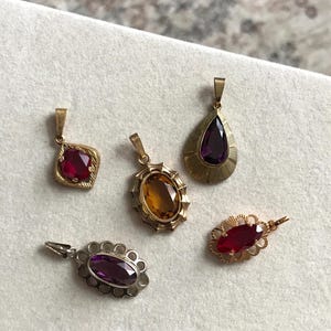 Puede incluir: Cinco colgantes de piedras preciosas vintage. Los colgantes presentan varias formas y colores, incluyendo piedras rojas, naranjas y moradas. Los colgantes están engastados en monturas de oro y plata, mostrando diseños y texturas intrincados. Una colección de joyas antiguas.
