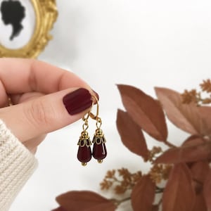 May include: A pair of drop earrings featuring deep burgundy, teardrop-shaped beads suspended from gold-toned metal findings. The beads are capped with intricate, scalloped gold-colored filigree accents, and the earrings are attached to gold-toned lever-back hooks.