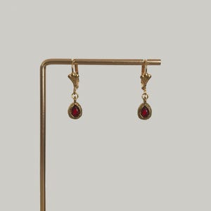 May include: A pair of gold-toned drop earrings featuring a small, teardrop-shaped red gemstone suspended from a lever-back closure. The earrings are displayed hanging from a thin, L-shaped gold-colored metal stand against a plain background.