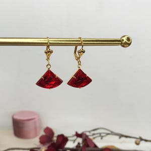 May include: A pair of gold-tone drop earrings featuring faceted, fan-shaped red crystals. Each earring is suspended from a gold-tone lever-back closure, showcasing a vibrant red gemstone cut into a wide, triangular fan silhouette with polished metal prongs.