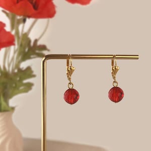 May include: A pair of gold-colored leverback earrings, each featuring a faceted, round, red bead. The earrings are displayed on a gold-colored stand, with red flowers in the background.