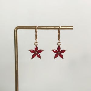May include: A pair of gold-toned leverback earrings featuring a five-petaled flower charm. Each flower is composed of five marquise-cut, deep red faceted stones arranged in a star shape, suspended from a simple gold-toned metal hoop mechanism.