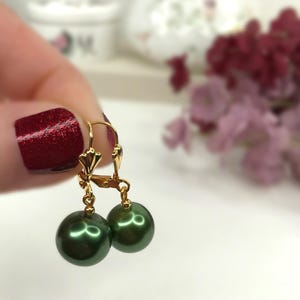 Può includere: Un paio di orecchini pendenti con perle rotonde verde scuro sospese a chiusure a monachella dorate. Le perle presentano una finitura liscia e lucente, mentre i componenti metallici mostrano una superficie color oro lucido con un piccolo dettaglio a ventaglio.