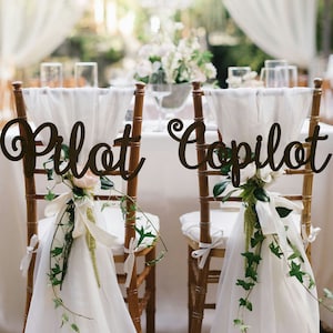 May include: Two white chairs with white fabric draped over the backs. The chairs are decorated with greenery and a white ribbon. A wooden sign on the back of each chair reads "Pilot" and "Copilot".