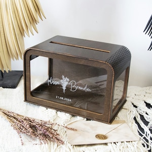May include: A wooden box with a clear acrylic lid and a slot for cards. The box is engraved with the names "Melissa & Brandon" and the date "21.08.2024".