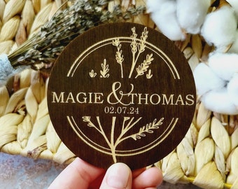 May include: A round wooden sign with a brown background and a light brown design. The sign features a floral design with the text "Maggie & Thomas 02.07.24".