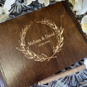 Op de afbeelding: Een donkerbruin houten gastenboek met een gouden gegraveerde krans en de tekst "Melissa & David 07.05.2024".