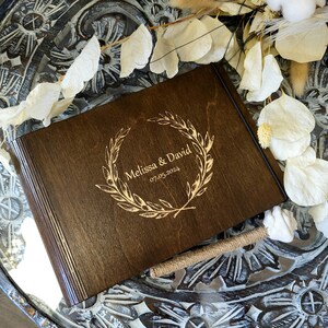 Op de afbeelding: Een donkerbruin houten gastenboek met een gouden gegraveerde krans en de tekst "Melissa & David 07.05.2024".
