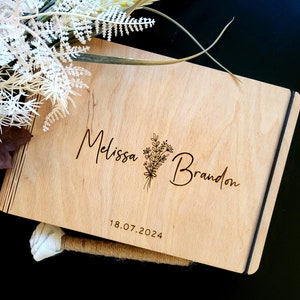 Op de afbeelding: Een houten gastenboek met een zwarte lint sluiting. De omslag is gegraveerd met de namen "Melissa" en "Brandon" en een bloemmotief. De datum "18.07.2024" is ook gegraveerd op de omslag.