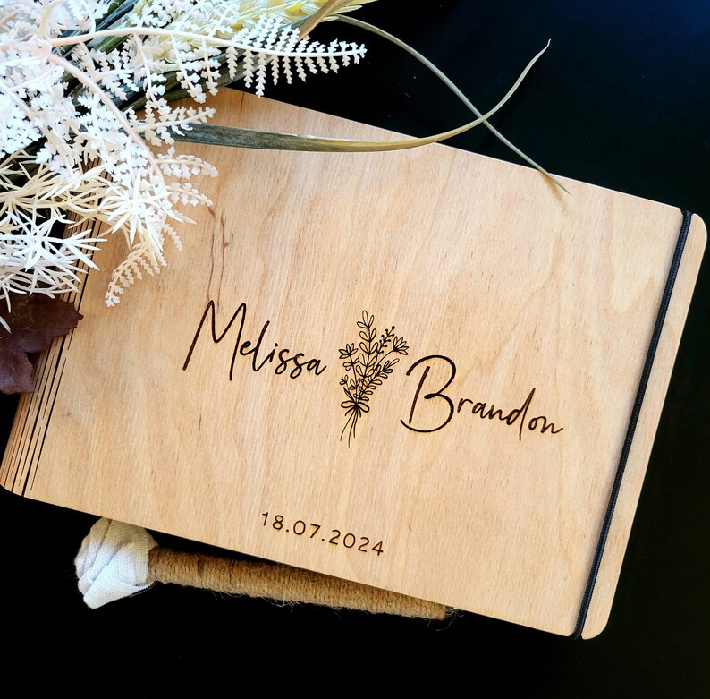 Op de afbeelding: Een houten gastenboek met een zwarte lint sluiting. De omslag is gegraveerd met de namen "Melissa" en "Brandon" en een bloemmotief. De datum "18.07.2024" is ook gegraveerd op de omslag.