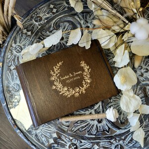 Op de afbeelding: Een donkerbruin houten gastenboek met de namen "Sophie & James" en de datum "05.07.2024" gegraveerd in een bloemenkrans. Het boek ligt op een decoratief oppervlak, met een met touw omwikkelde pen.