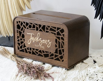 Puede incluir: Una caja de madera marrón oscuro con un diseño de corte geométrico y una ranura en la parte superior. La caja tiene una inscripción dorada que dice "Jacksons 12.07.2025".