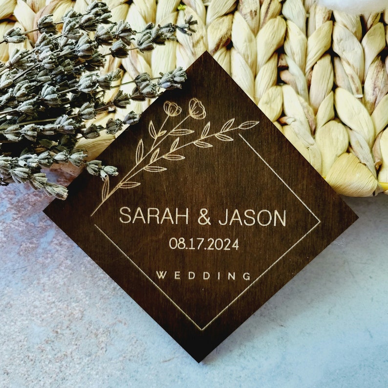 May include: A dark brown wooden square sign with a gold floral design and the text "SARAH & JASON 08.17.2024 WEDDING" in a white square frame.