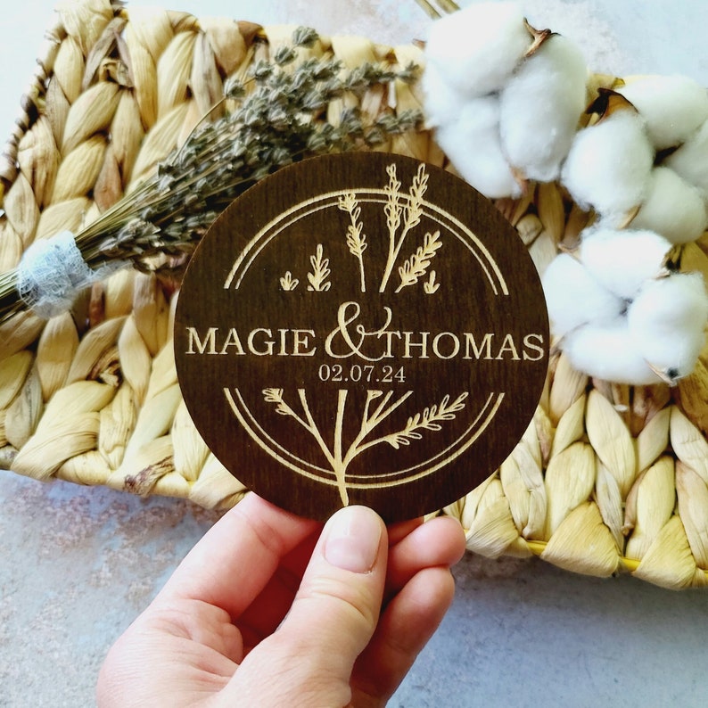 May include: A round wooden sign with a brown background and a light brown design. The sign features a floral design with the text "Maggie & Thomas 02.07.24".