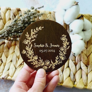 Può includere: Un sottobicchiere rotondo in legno con un design floreale inciso in un colore marrone chiaro. Il sottobicchiere ha il testo "Sophie & James" e la data "05.07.2024" incisi su di esso.