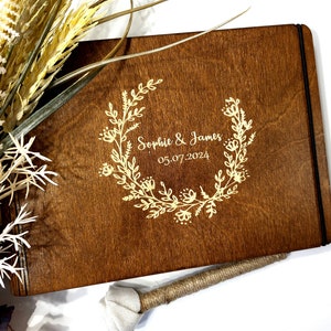 Op de afbeelding: Een houten gastenboek met een zwarte lint sluiting. De omslag is gegraveerd met een krans van bloemen en de tekst "Sophie & James 05.07.2024".