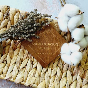 May include: A wooden sign with a diamond shape and gold lettering. The sign says "Sarah & Jason" and "08.17.2024" with the word "Wedding" below the date. The sign is sitting on a woven straw mat with dried lavender and cotton balls.
