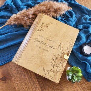May include: A rectangular wooden photo album with a light brown finish, featuring laser-engraved floral illustrations and the text "WHERE OUR STORY LIVES Samantha & Jonathan est. May 18th, 2027". A small gold-coloured heart-shaped padlock secures the cover.